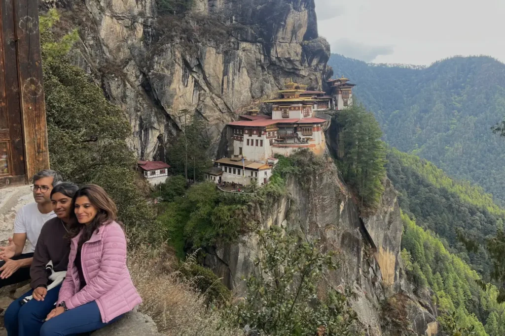 A family sits together, smiling for a photo during their trek, with the iconic Tiger’s Nest Monastery perched dramatically on the cliffside in the background, surrounded by lush green mountains