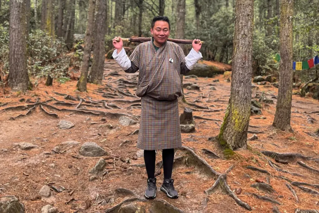 A Bhutanese guide in traditional attire (Gho) standing on a forest trail during the hike to Tiger’s Nest Monastery, surrounded by tall pine trees and exposed tree roots.