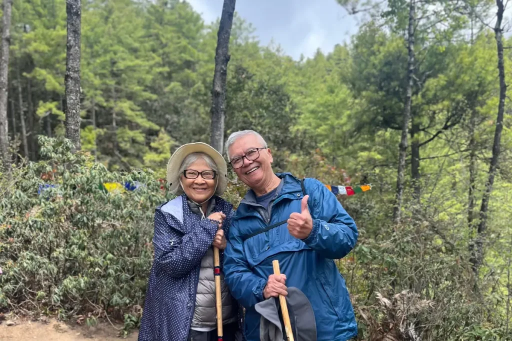 Phil and Cookie from Dallas, smiling during a forest trek with walking sticks and prayer flags in the background.