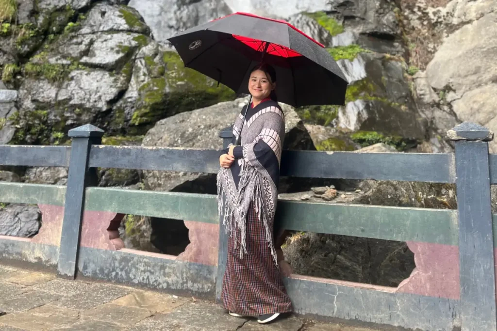 A pretty Bhutanese woman stands in front of a waterfall, wearing traditional dress