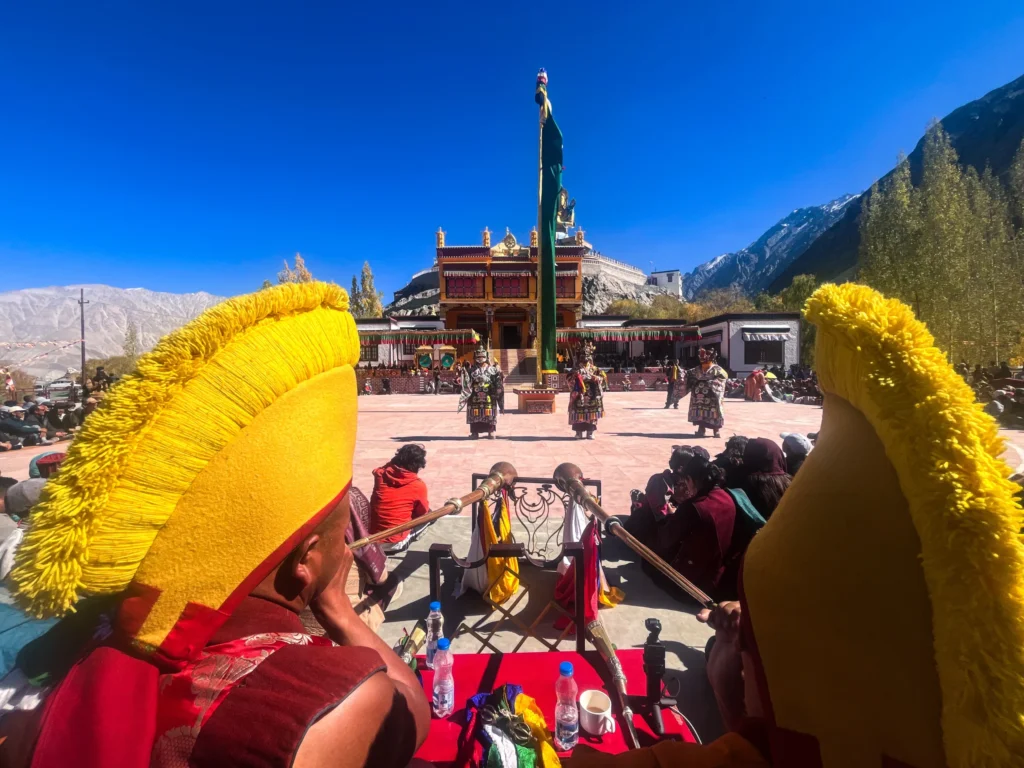 Monks wearing traditional yellow hats observe the Diskit Gustor Festival at Diskit Monastery in Ladakh, with masked dancers performing against a backdrop of clear blue skies and Himalayan mountains